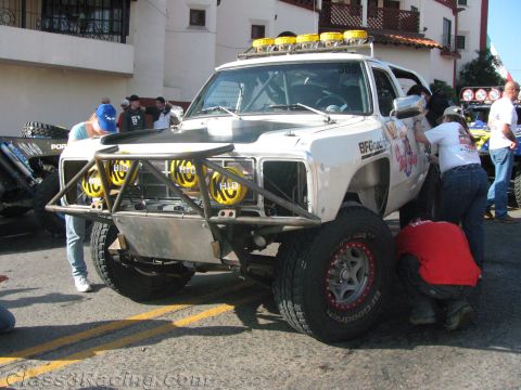 1990 Ford Bronco - white off-road race truck with a black hood and yellow-lensed lights, crew members working around it in a staging area.