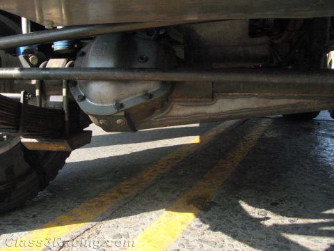1990 Ford Bronco - Low underbody view of an off-road truck showing the front differential, solid axle and leaf spring suspension over a painted street.