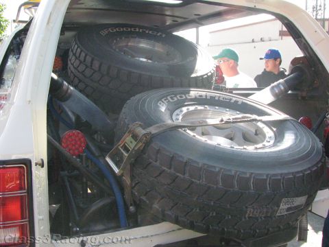 1990 Ford Bronco - View into the open rear of a white off-road race SUV stacked with three BFGoodrich spare tires as two men look on.