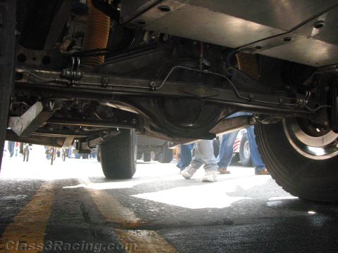 1990 Ford Bronco - Low underbody view of an off-road vehicle showing the rear axle and coil-spring suspension with spectators' legs visible behind.