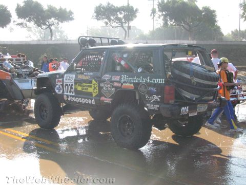 1990 Ford Bronco - black lifted Jeep Cherokee race vehicle, number 1705, with roof light bar and sponsor decals stands on wet pavement among other off-road ri…