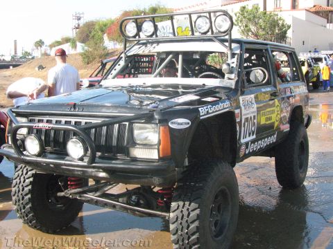 1990 Ford Bronco - black lifted Jeep Cherokee race rig, number 1705, shows its grille, roof light bar and coilover suspension on a wet paved staging area.