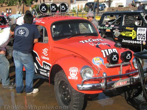 1990 Ford Bronco - red Volkswagen Beetle off-road race car, number 1100, with twin auxiliary lights and Tecate decals while people work beside it.