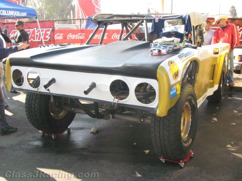 1990 Ford Bronco - yellow and white tube-chassis off-road race vehicle sits on jack stands in a pit area surrounded by Tecate and Coca-Cola signage.