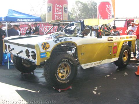 1990 Ford Bronco - yellow and white tube-chassis off-road race vehicle on red jack stands in a pit area, marked with BFGoodrich and KC decals.