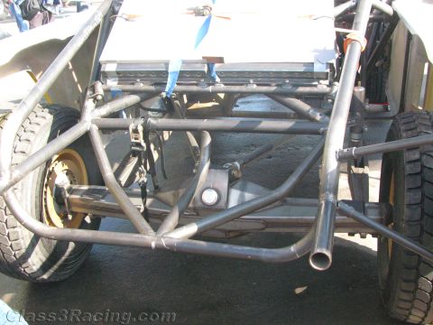 1990 Ford Bronco - underside of an off-road race vehicle showing its rear tube chassis, suspension links and exhaust pipe on paved ground.