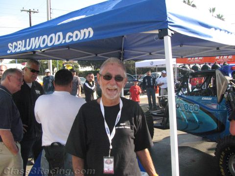 1990 Ford Bronco - smiling gray-bearded man in sunglasses and a black shirt stands under a blue SCBLUWOOD.com canopy with a teal race vehicle behind him.