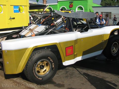 1990 Ford Bronco - white and gold open tube-frame off-road race vehicle with a rear wing and BFGoodrich decals parked in a pit near a yellow trailer.