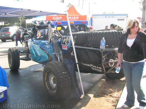 1990 Ford Bronco - teal open-frame off-road race buggy, number 124, with rear spare tires sits in a pit area as a woman walks past holding a water bottle.
