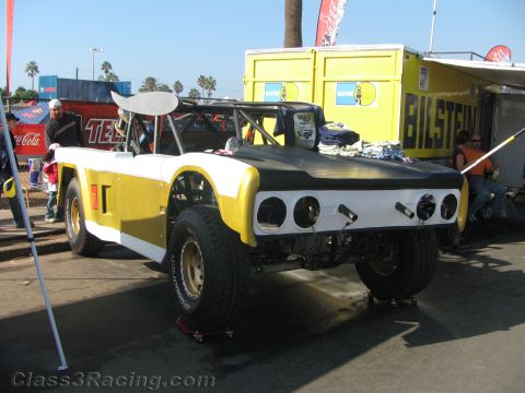 1990 Ford Bronco - yellow and white open tube-frame off-road race vehicle with a rear wing sits on jack stands beside a yellow Bilstein trailer in a pit.