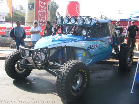 1990 Ford Bronco - blue BLUWOOD-branded open-frame off-road race vehicle with roof light bar and big tires draws onlookers in a pit area.