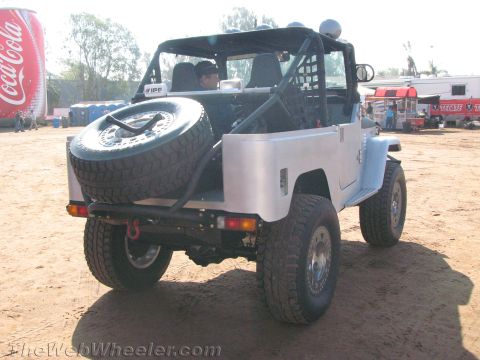 1990 Ford Bronco - silver bare-metal open-top FJ-style 4x4 with a rear-mounted spare tire and roll cage parked on dirt at an event.