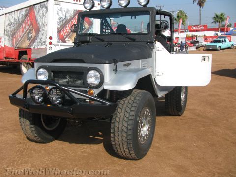1990 Ford Bronco - silver and black FJ-style 4x4 with a tubular brush bar, auxiliary lights and roof light bar parked on dirt near Tecate trailers.