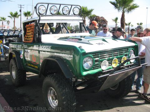 1990 Ford Bronco - green and white early Ford Bronco race truck, number 301, with roof light bar and brush bar surrounded by a crowd at an event.