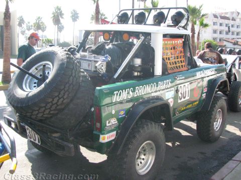 1990 Ford Bronco - green early Ford Bronco race truck, number 301, with rear spare tire and Tom's Bronco Parts lettering parked among palm trees.
