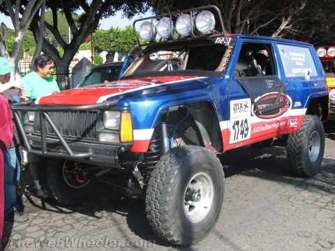 1990 Ford Bronco - blue, red and white lifted Jeep Cherokee race vehicle, number 1749, with roof light bar and brush bar parked on a street near trees.