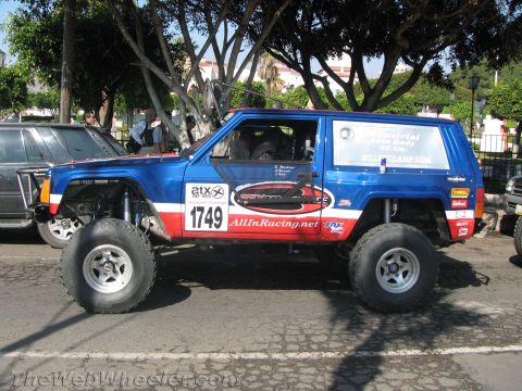 1990 Ford Bronco - side profile of a blue, red and white lifted Jeep Cherokee race vehicle, number 1749, with AllInRacing.net decals on a tree-lined street.