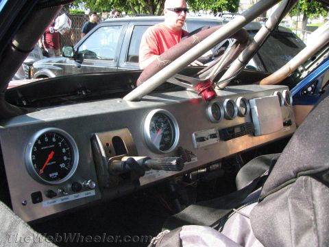 1990 Ford Bronco - Bare off-road race-truck cockpit showing a roll cage, large analog tachometer and a row of toggle switches across a metal dash.