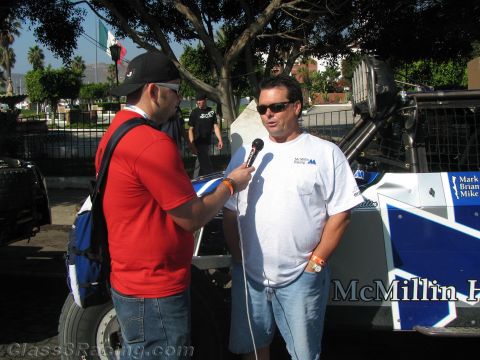 1990 Ford Bronco - man in a white McMillin Racing shirt and sunglasses is interviewed with a microphone beside a blue race vehicle outdoors.