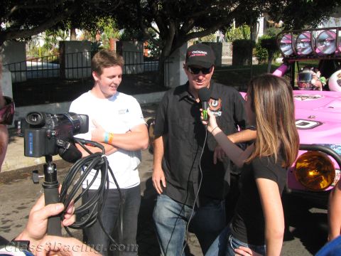1990 Ford Bronco - camera operator films two men being interviewed by a woman with a microphone next to a pink off-road race truck.