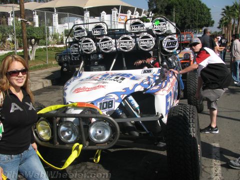 1990 Ford Bronco - white-and-blue number 108 off-road buggy with a Hella roof light bar parked on a street as a woman stands at the front.