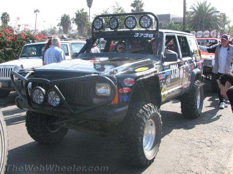 1990 Ford Bronco - lifted black Jeep Cherokee race rig numbered 3736, fitted with KC lights and a roof light bar, parked among other off-roaders.