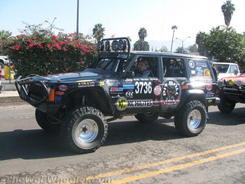 1990 Ford Bronco - black lifted Jeep Cherokee race truck numbered 3736 with KC Hilites and sponsor decals drives along a sunny street.