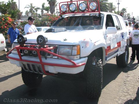 1990 Ford Bronco - white lifted SUV race rig numbered 302 with a red front tube bumper, driving lights and a roof light bar at an event.
