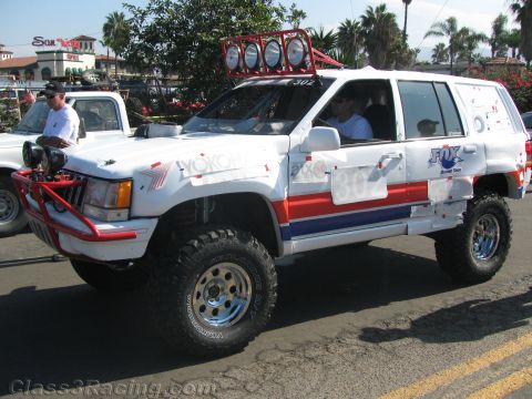 1990 Ford Bronco - white SUV racer with red and blue stripes, number 302 and a roof light bar drives down a street lined with palms.