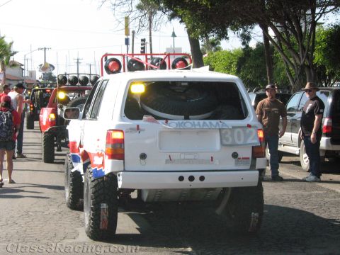 1990 Ford Bronco - Rear view of a white SUV racer number 302 carrying a spare tire, queued in a street line of off-road vehicles with onlookers.
