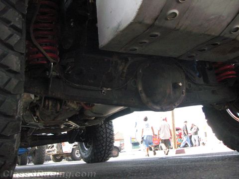 1990 Ford Bronco - Underbody view of a lifted off-road vehicle revealing red coilover shocks, the rear axle housing and large mud tires.