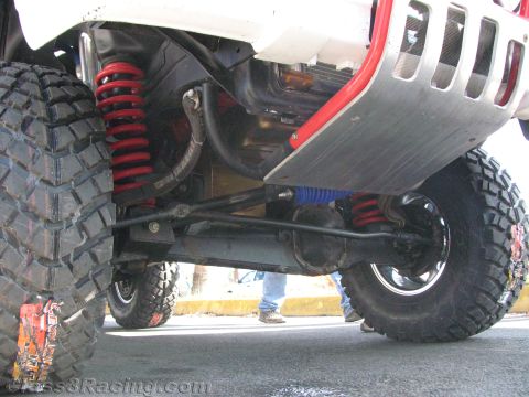 1990 Ford Bronco - Underside of a red off-road vehicle showing red and blue coilover shocks, a skid plate and chunky mud-terrain tires.