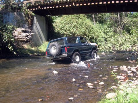 1979 Ford Bronco - A dark blue SUV navigates a shallow river, with a wooden bridge overhead and lush greenery surrounding the banks.