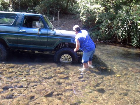 1979 Ford Bronco - A man wades in a stream next to a green 1979 Ford Bronco as it drives through the water.