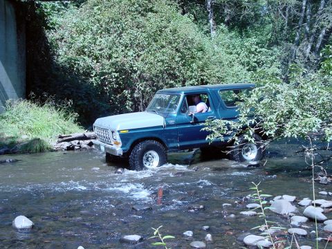 1979 Ford Bronco - A blue and silver 1979 Ford Bronco navigates a rocky stream bed surrounded by lush greenery.