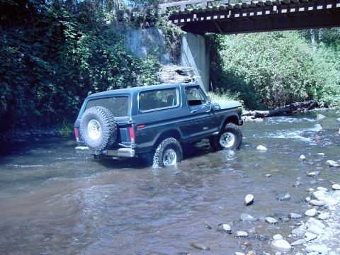 1979 Ford Bronco - Ford Bronco navigates a rocky stream bed beneath a bridge, its tires partially submerged in water.