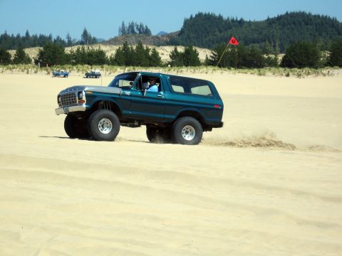 1979 Ford Bronco - A lifted Ford Bronco kicks up sand while driving on a dune, with other vehicles visible in the distance.