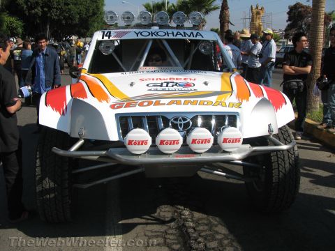 1990 Ford Bronco - Front view of a white number 14 Toyota off-road race truck with Geolandar and Koito lights and a roof light bar on a street.
