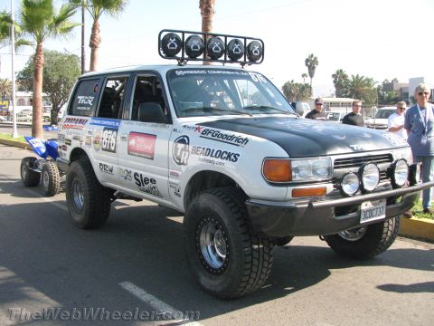 1990 Ford Bronco - white-and-black Toyota Land Cruiser race rig numbered 676 with a roof light bar tows a blue ATV on a trailer at an event.