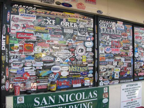 1990 Ford Bronco - storefront window densely covered with racing and brand stickers above a green San Nicolas parking sign.