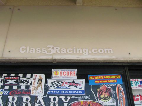 1990 Ford Bronco - shop window plastered with off-road racing stickers including SCORE, All Valley Carburetor and Pro-Racing decals beneath a watermark.