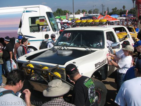1990 Ford Bronco - white pre-running truck with a black hood scoop and BFGoodrich lettering draws a crowd of spectators at an off-road racing gathering.