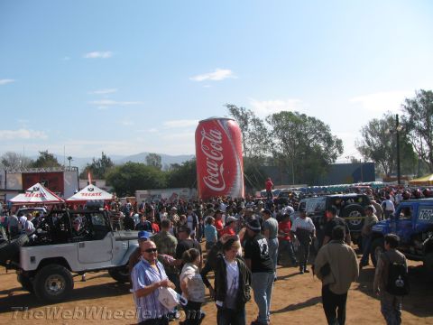 1990 Ford Bronco - large crowd gathers on a dirt lot beside off-road vehicles, a giant inflatable Coca-Cola can and Tecate tents under a clear sky.