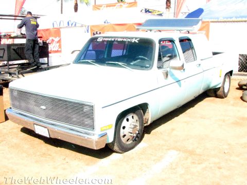 1990 Ford Bronco - lowered light-blue Chevrolet crew-cab dually pickup with a billet grille and Streetshock banner sits on a dirt lot at an event.