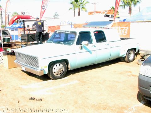 1990 Ford Bronco - lowered light-blue Chevrolet crew-cab dually pickup sits on a dirt lot near event tents, banners and palm trees.