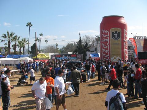 1990 Ford Bronco - Spectators pack a dirt street beneath a towering inflatable Tecate can near start and finish banners at an off-road racing event.