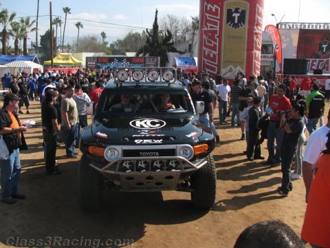 1990 Ford Bronco - black Toyota FJ Cruiser race vehicle with KC roof lights and a tubular bumper is surrounded by spectators near a Tecate inflatable.