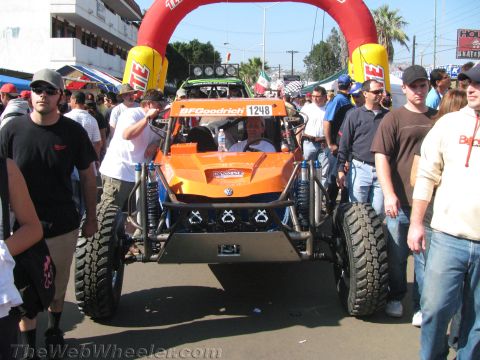 1990 Ford Bronco - orange tube-frame off-road buggy numbered 1248 with BFGoodrich tires waits under a red start arch amid a packed crowd.