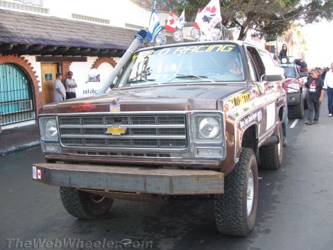 1990 Ford Bronco - brown 1970s-era Chevrolet Blazer prerunner with a tall snorkel, roof lights and Yolo Racing banner moves through a crowded street.