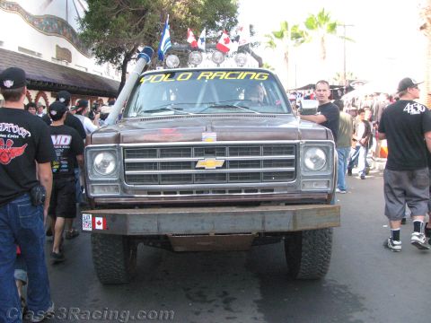 1990 Ford Bronco - brown Chevrolet prerunner with a Yolo Racing windshield banner, roof lights and snorkel sits among black-shirted spectators.